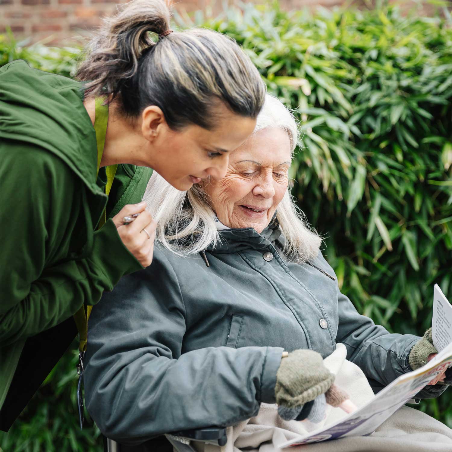 A younger woman smiles with an elderly woman in a wheelchair, looking at a magazine outdoors. - Home Instead