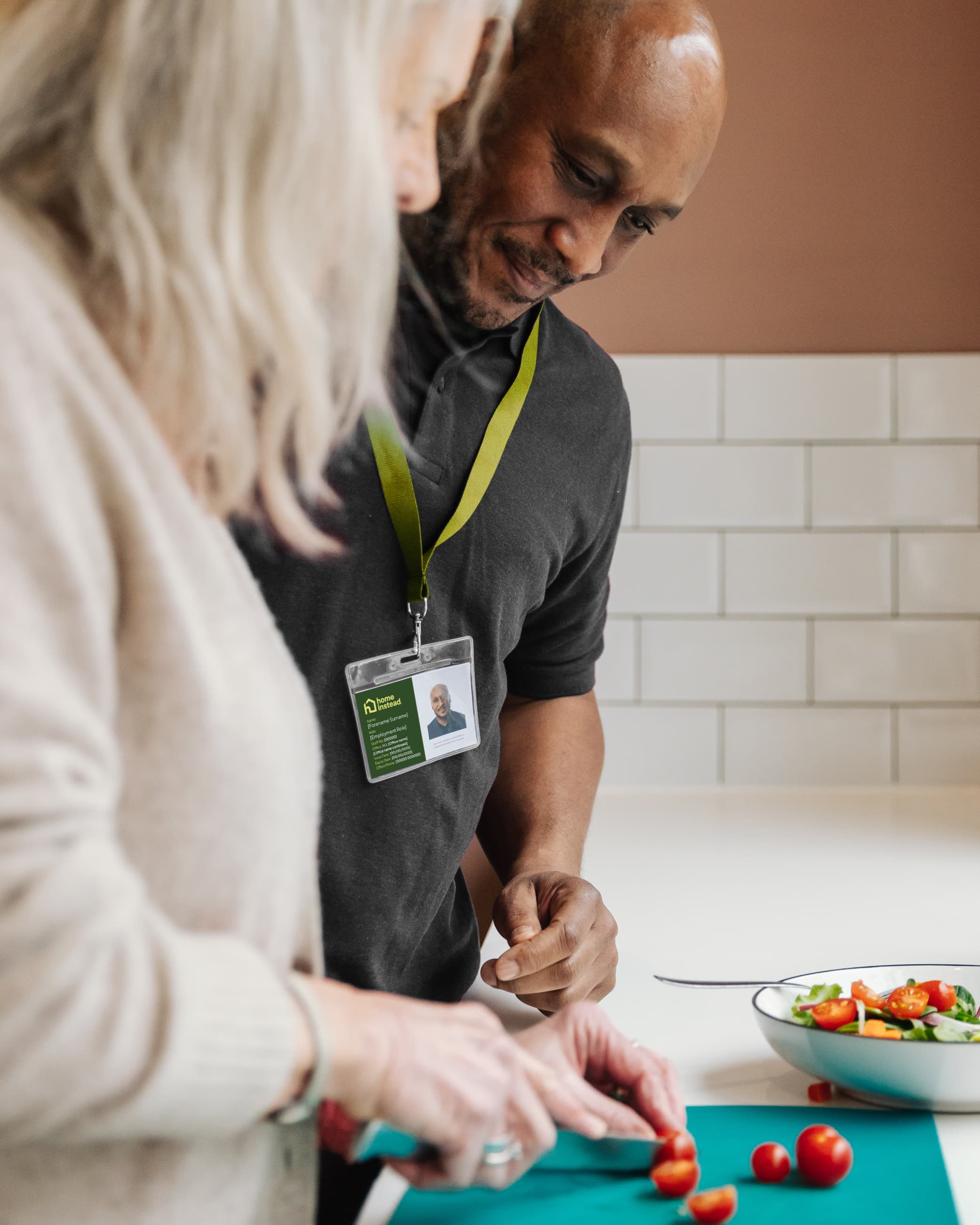 Two people slice cherry tomatoes together in a kitchen; one wears a green lanyard with an ID badge. - Home Instead
