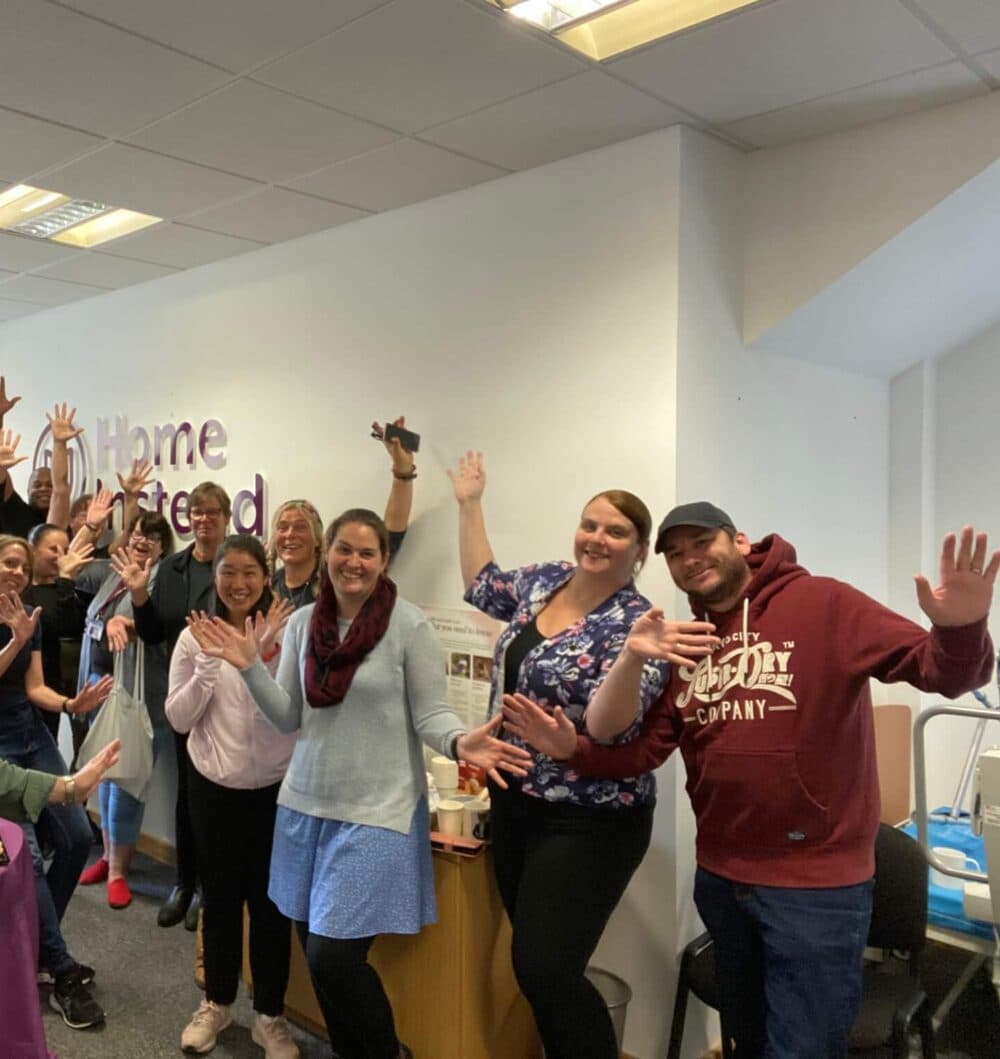 Group of people smiling and waving in an office setting with "Home Instead" sign in the background. - Home Instead