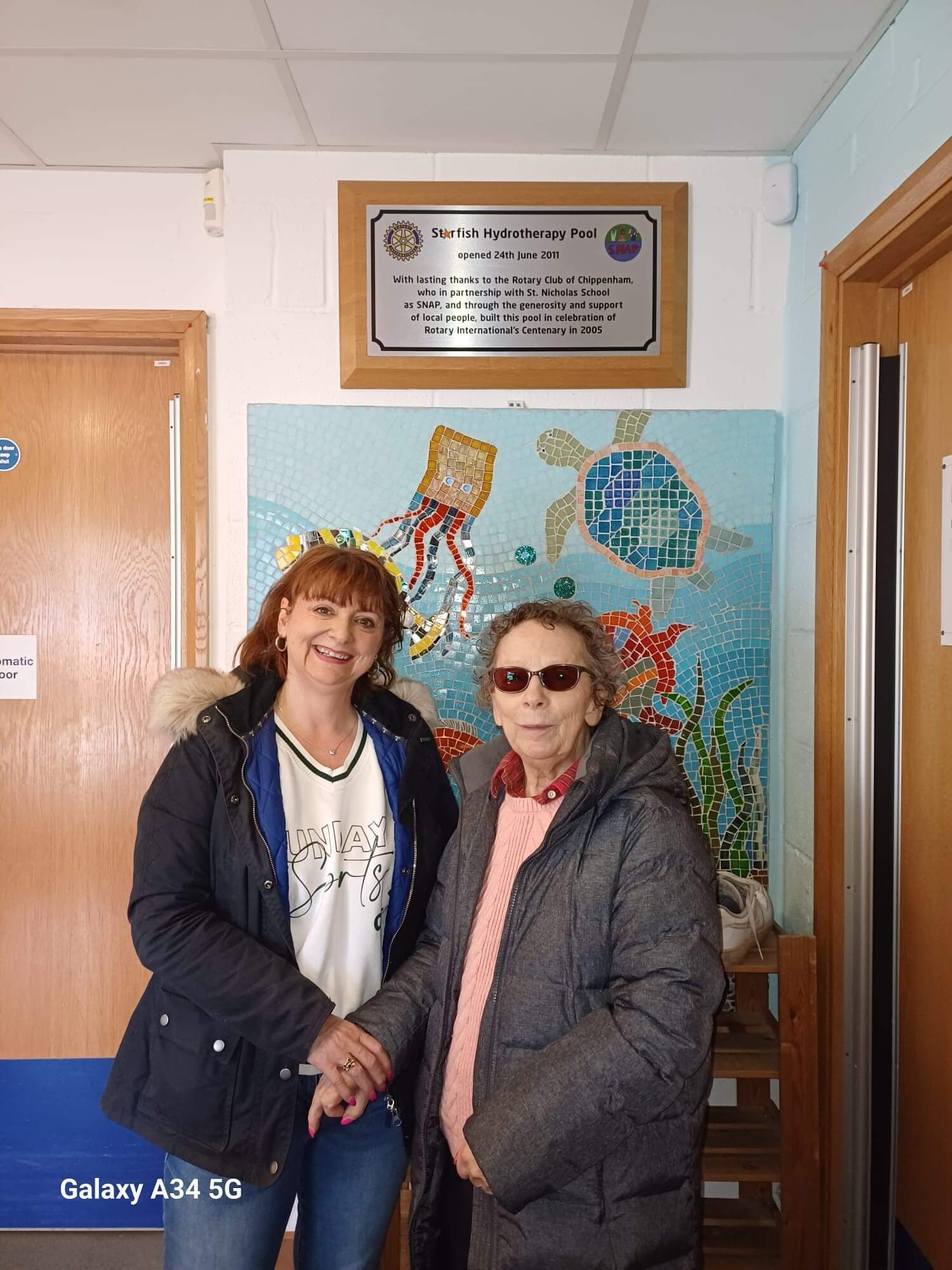 Two women smiling and holding hands in front of a colorful mural and a plaque in a building lobby. - Home Instead