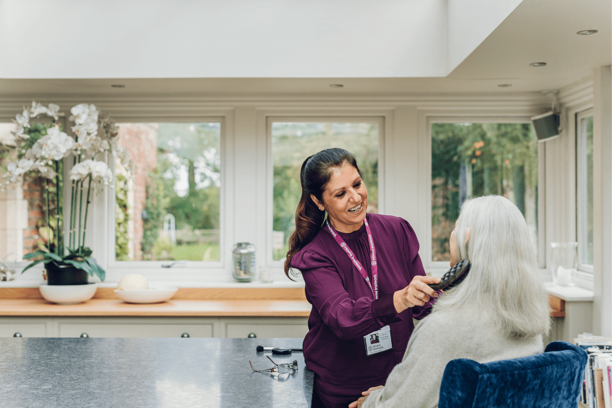 A caregiver in a purple top brushes an elderly woman's hair in a bright, sunlit room. - Home Instead