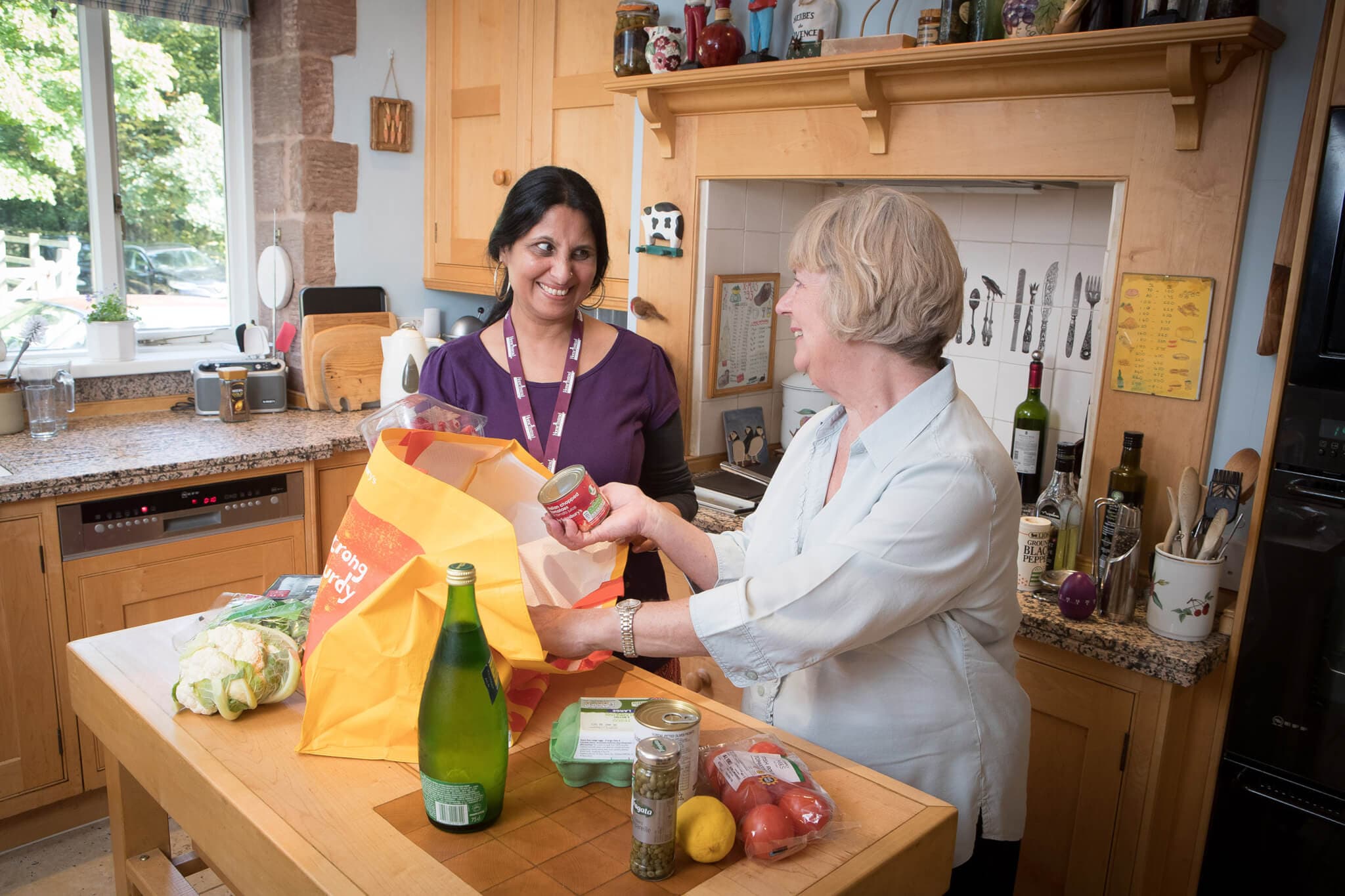 Two women unpack groceries in a kitchen, smiling at each other. One is wearing a name badge and the other is holding a can. - Home Instead