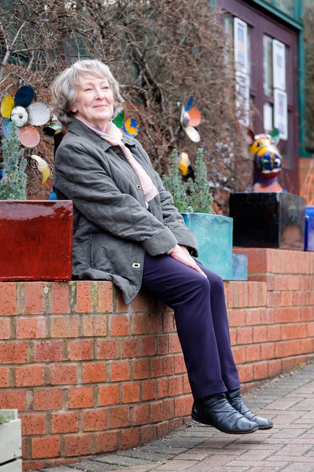 Elderly woman with short hair, wearing a coat and boots, sitting on a brick planter outdoors, smiling. - Home Instead