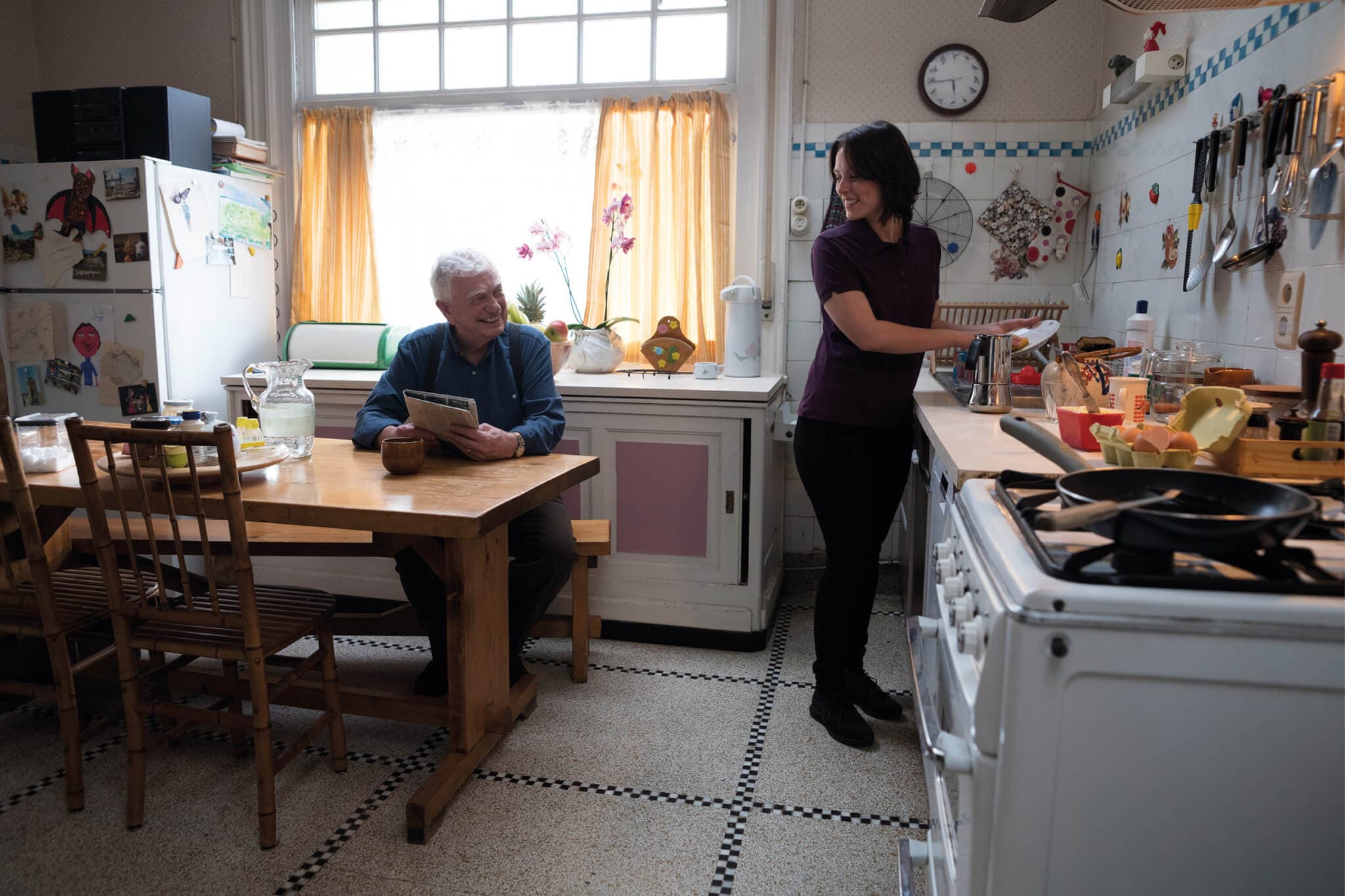 An older man reads a newspaper at a kitchen table while a younger woman prepares food at a counter. - Home Instead
