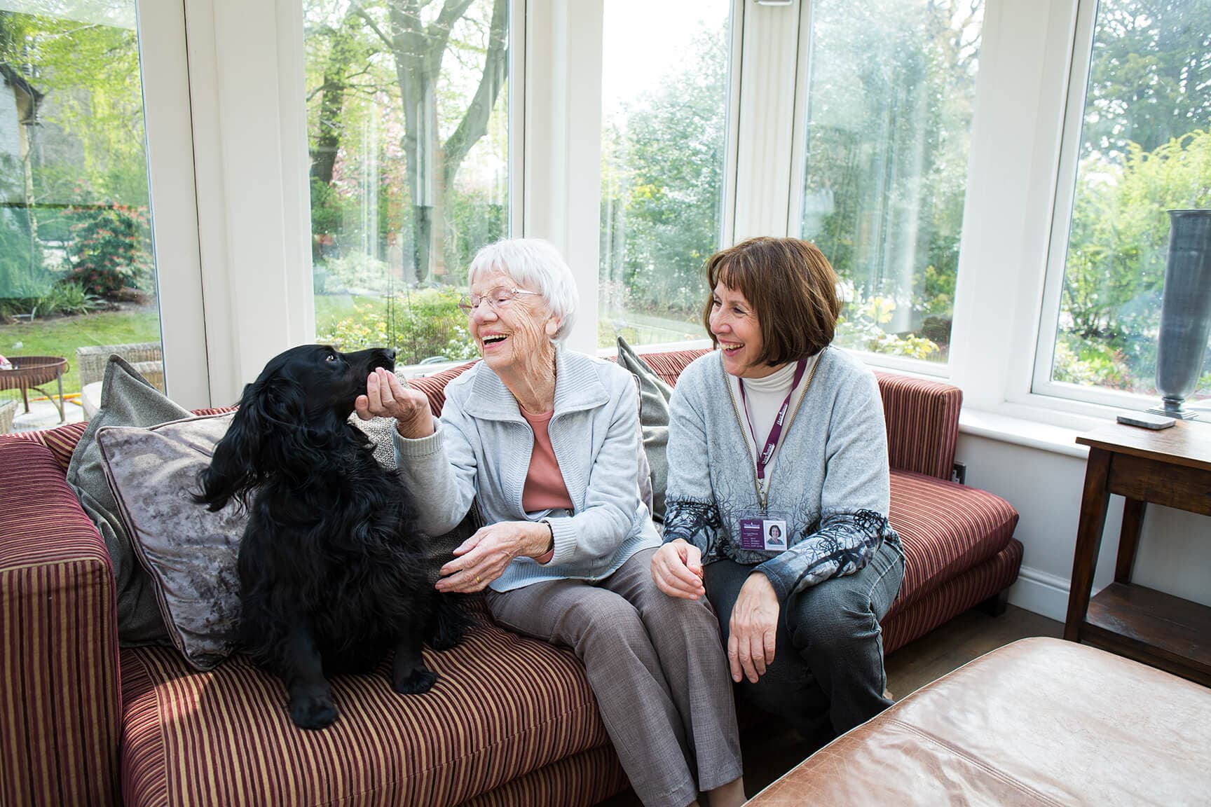 An elderly woman pets a black dog while sitting on a couch with another woman in a sunlit room. - Home Instead