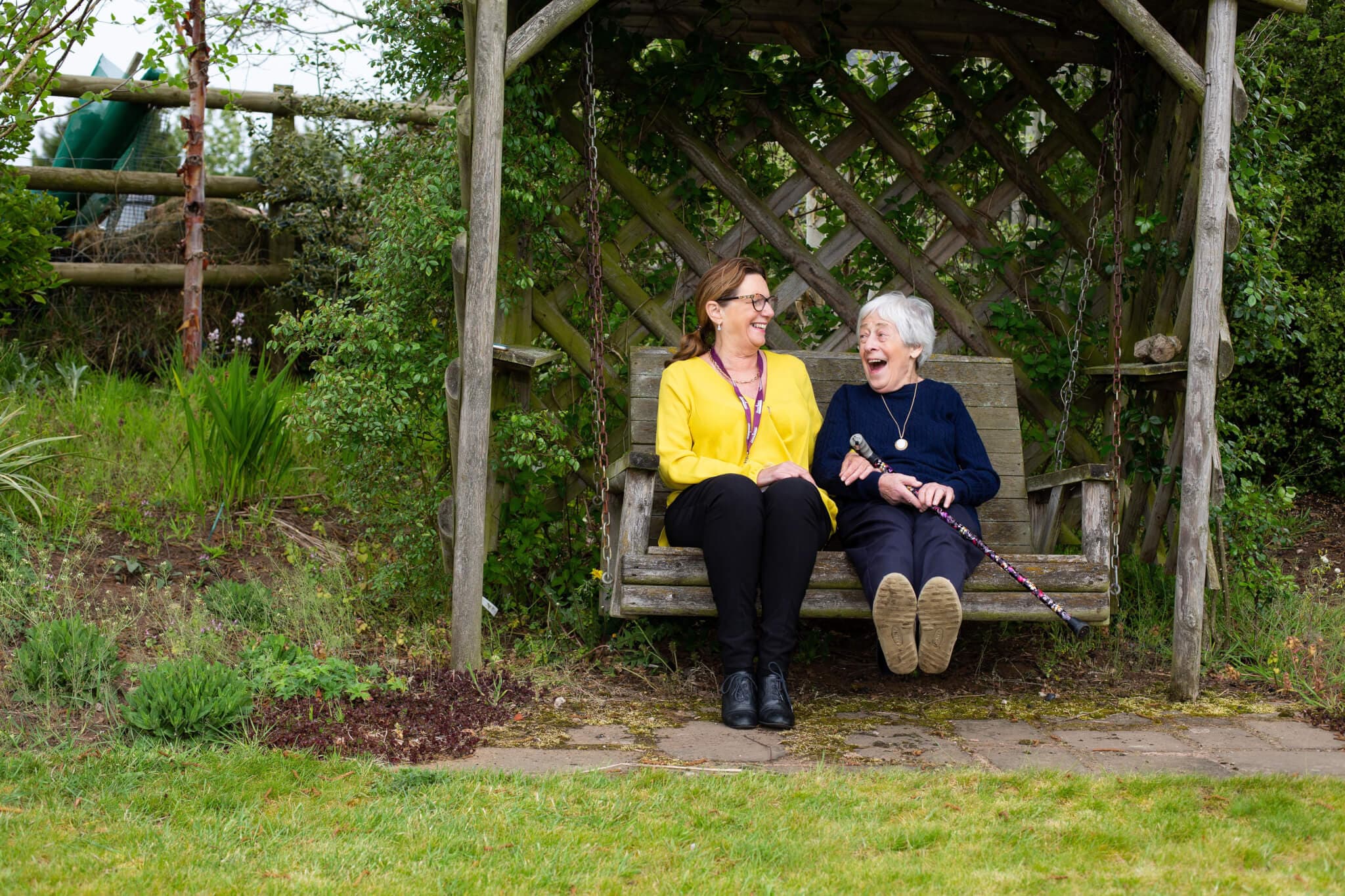 Two women sitting on a garden swing, smiling and talking, surrounded by greenery and wooden trellis. - Home Instead