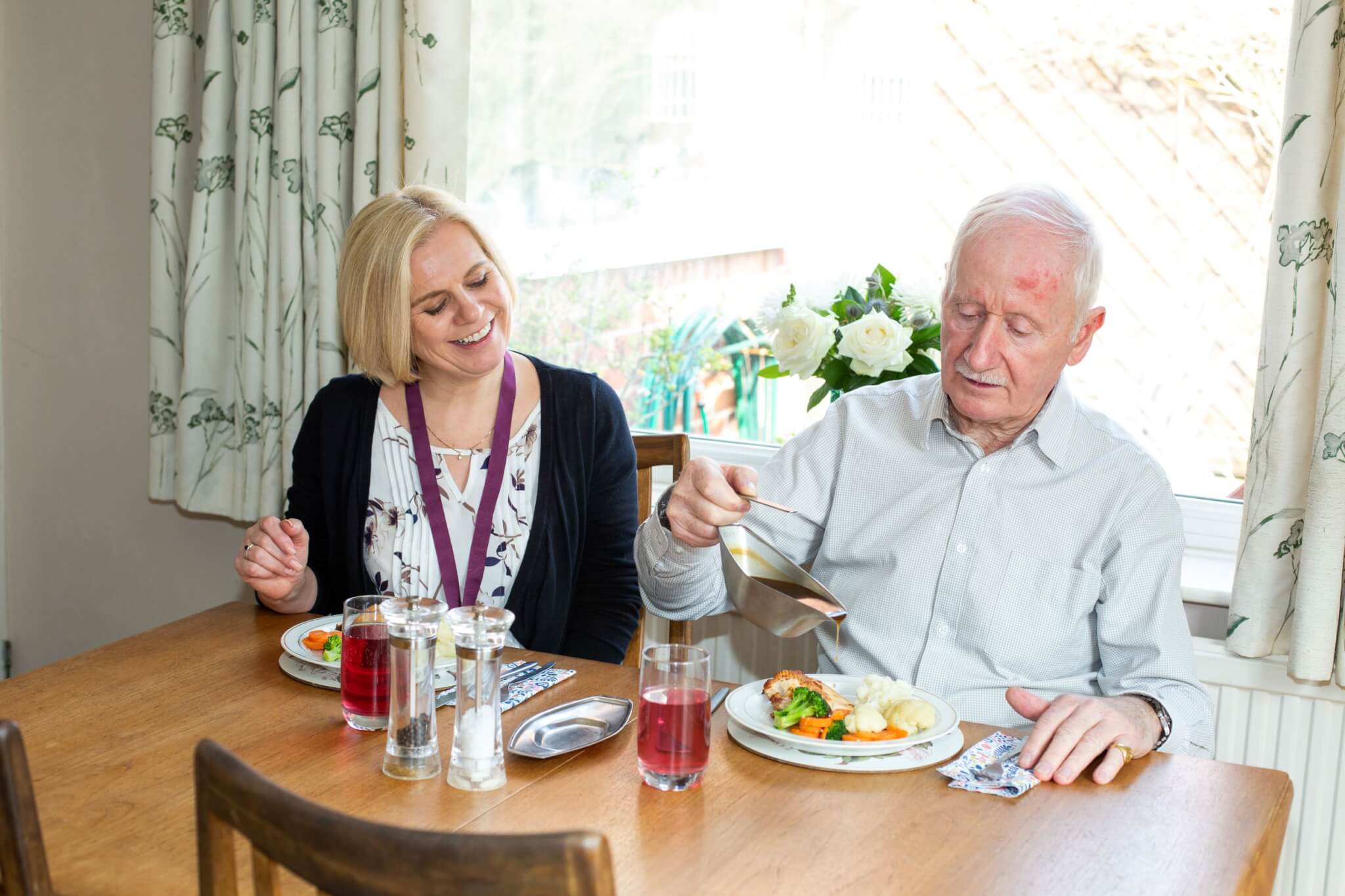 A woman and an elderly man share a meal at a wooden dining table, with flowers and drinks visible. - Home Instead