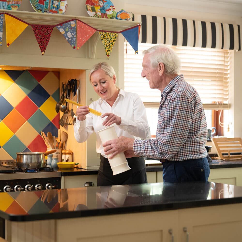 A woman and an elderly man are in a kitchen preparing food together, with colorful bunting in the background. - Home Instead