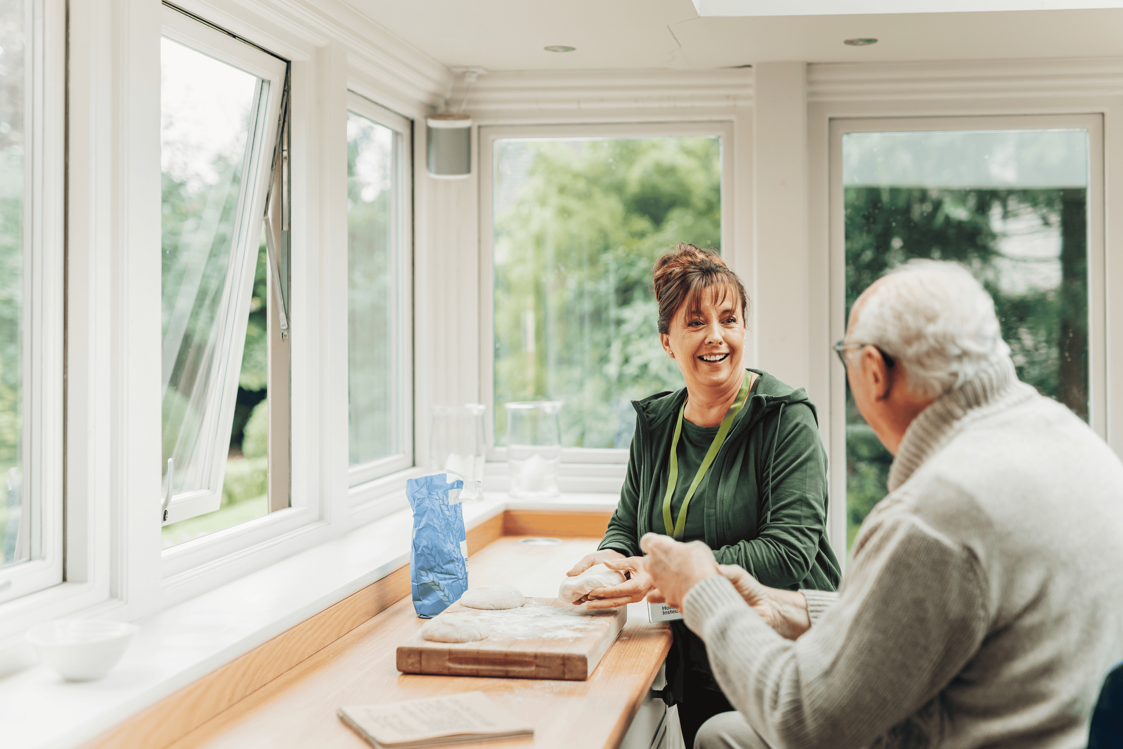 Two people sit by a bright window, smiling and preparing dough together on a wooden countertop. - Home Instead