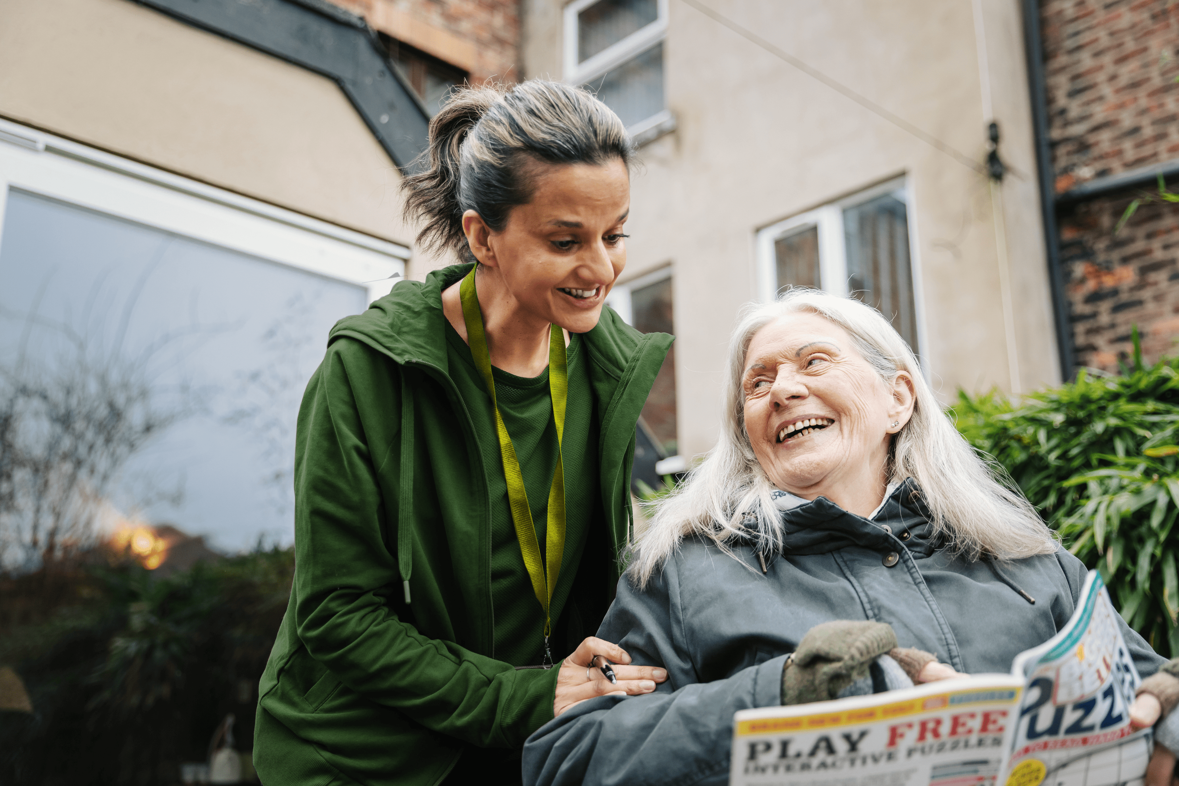 A young woman and an older woman smile together outdoors, holding a puzzle magazine. - Home Instead