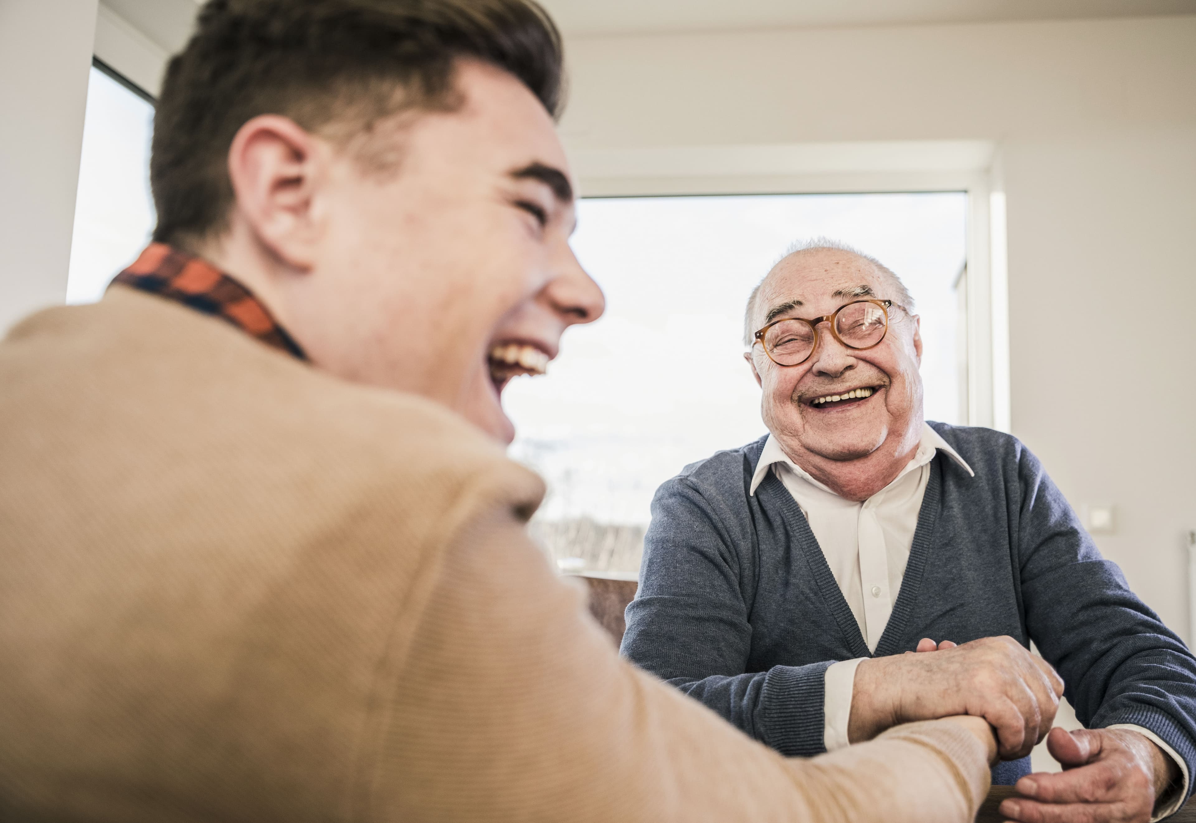 A young man and an elderly man laughing together indoors, sitting by a window. - Home Instead