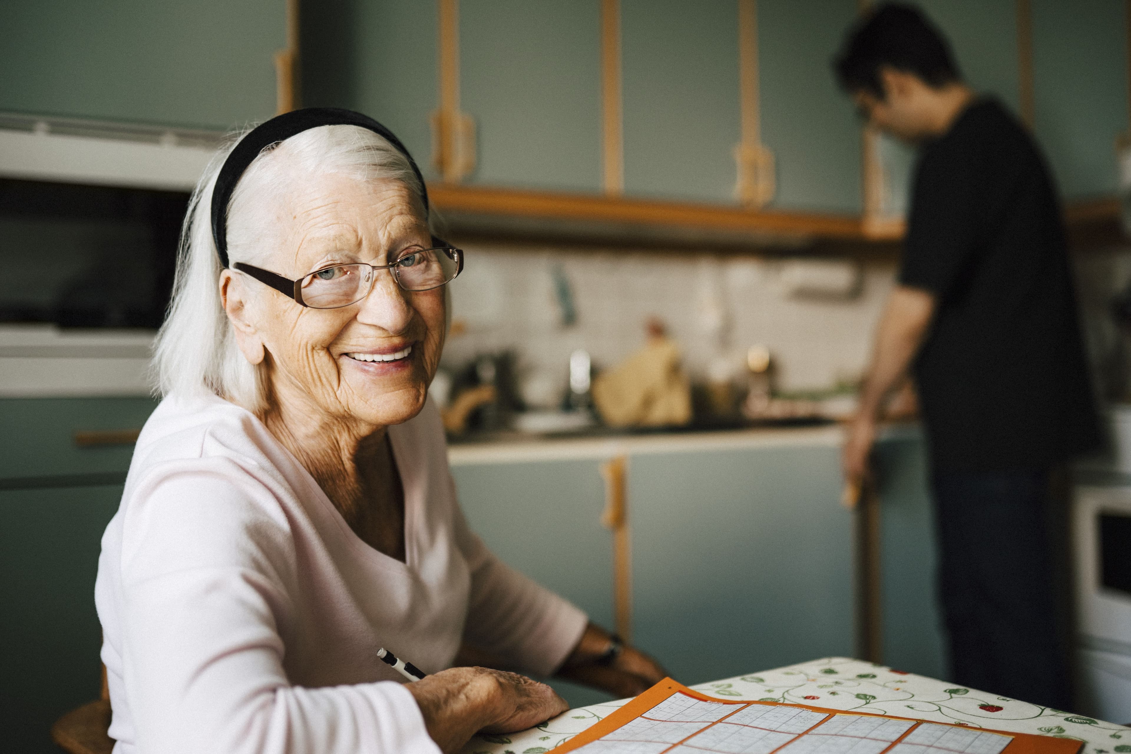 Smiling elderly woman with glasses sits at a table while a man stands in the kitchen in the background. - Home Instead