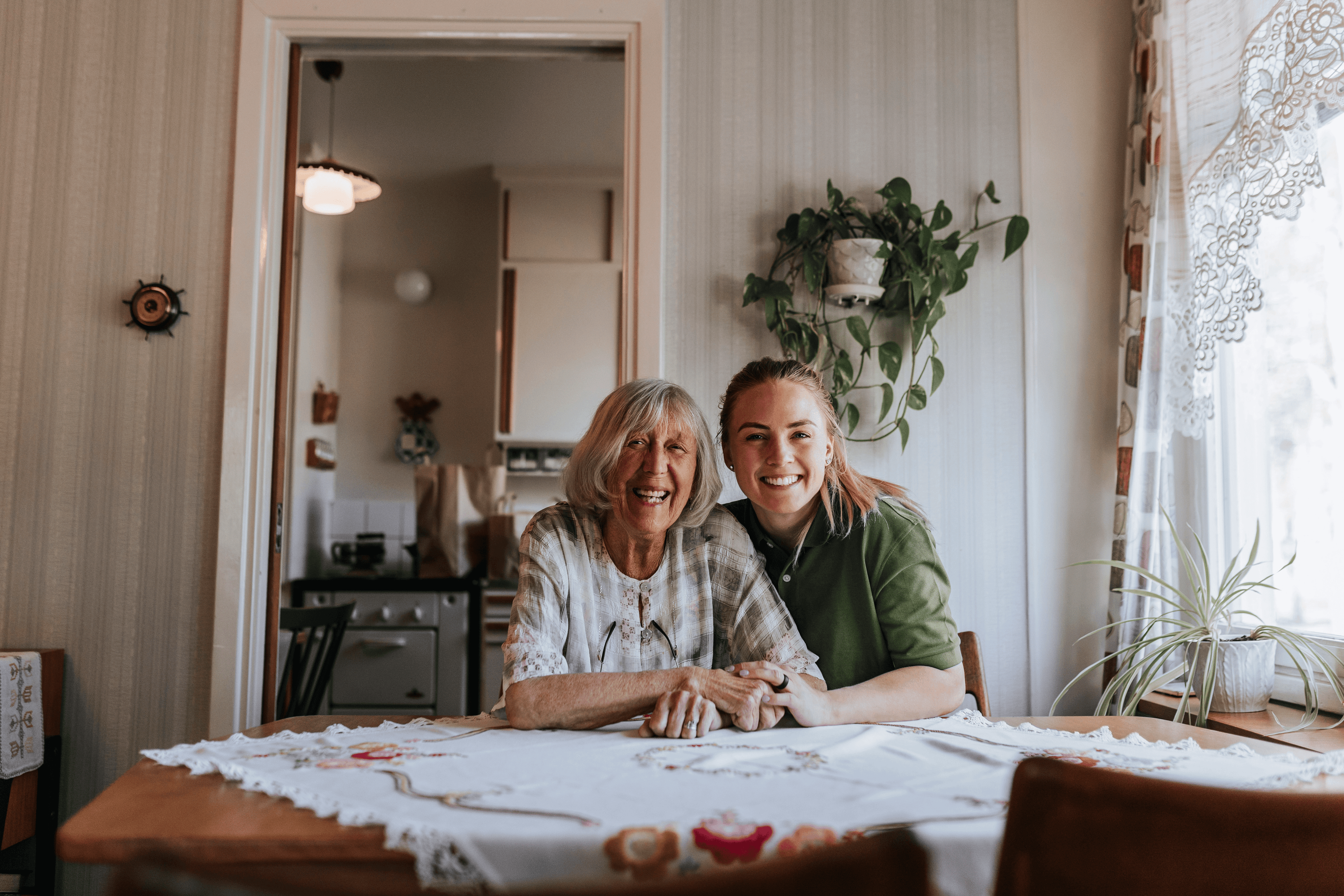 Two women smiling and sitting at a table in a cozy, sunlit kitchen with plants and lace curtains. - Home Instead