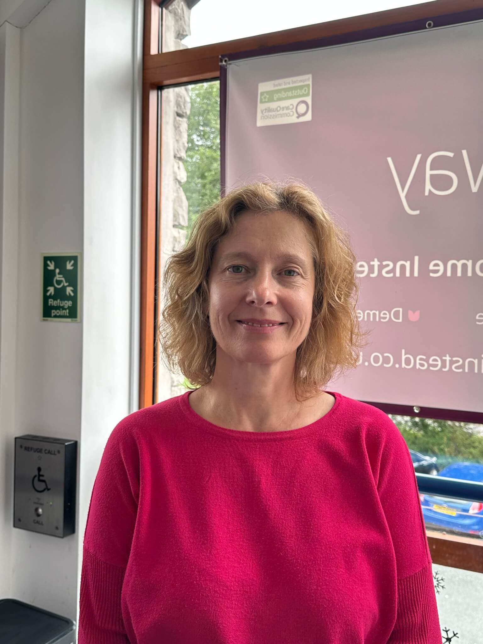 A person with short curly hair wearing a bright pink top stands indoors, smiling in front of a window. - Home Instead