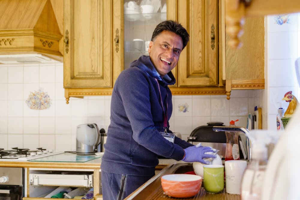 A smiling person in a blue hoodie and gloves washes dishes in a cozy, wooden-themed kitchen. - Home Instead