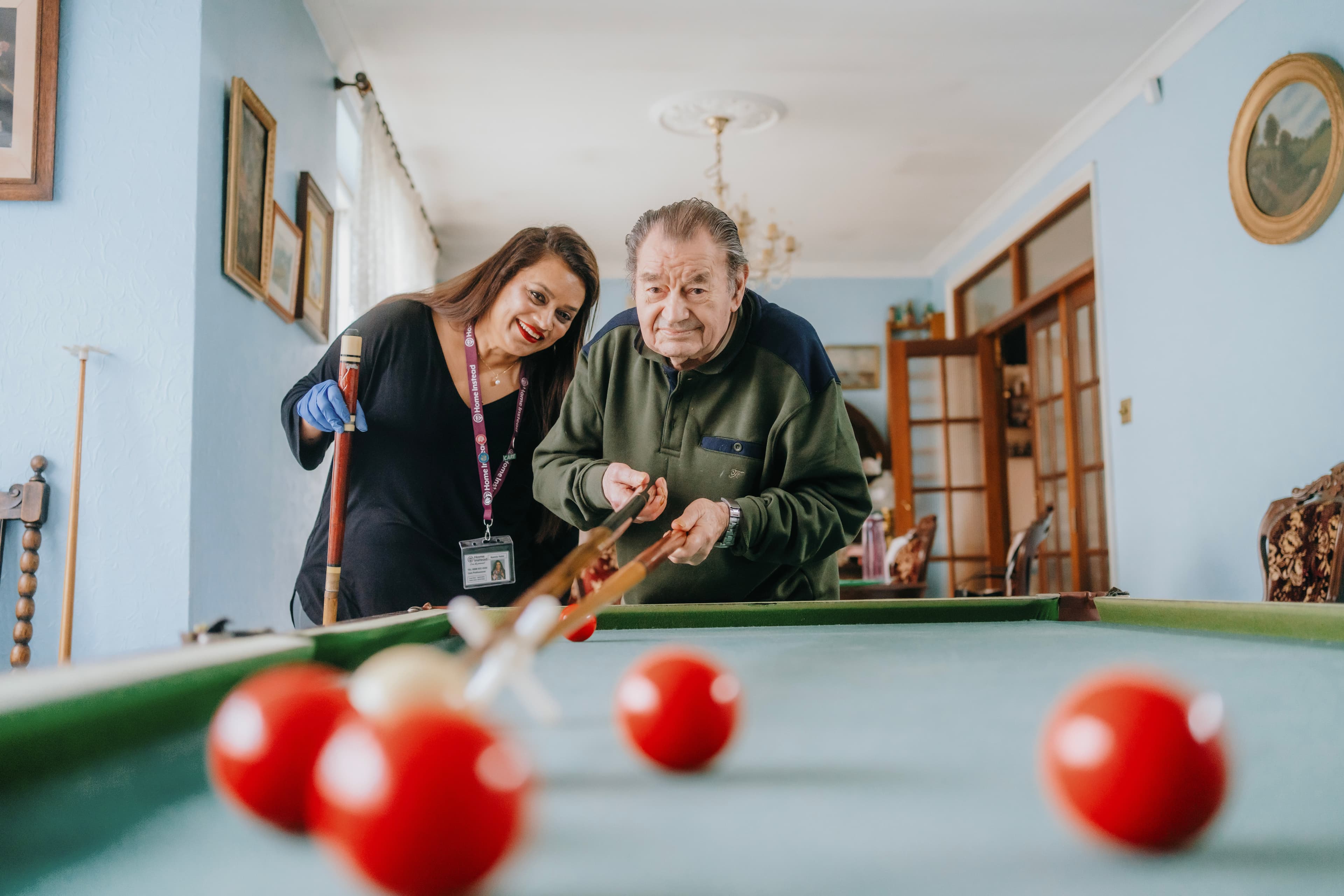Elderly man playing pool, assisted by a smiling woman in a home setting. Red balls are on the table. - Home Instead