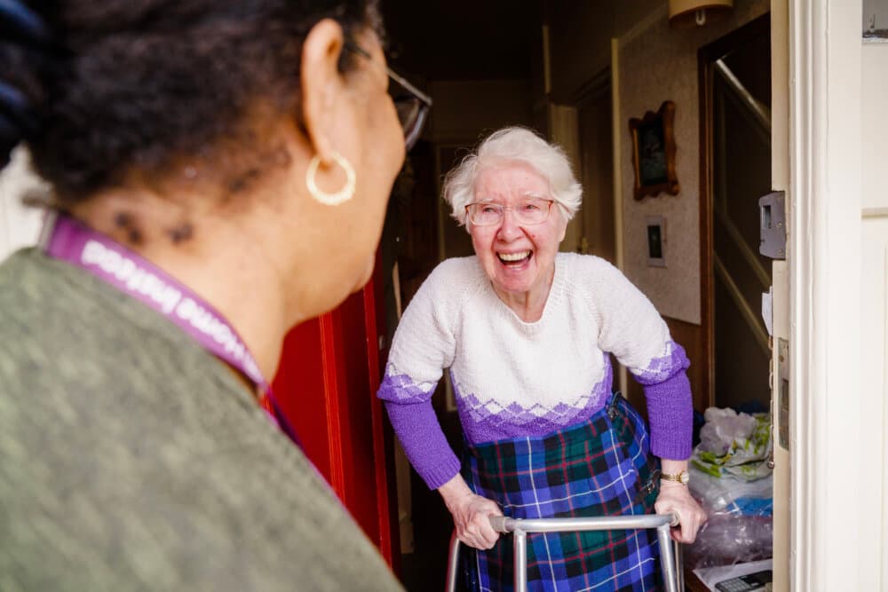 Elderly woman using a walker smiles at another woman at her door. - Home Instead