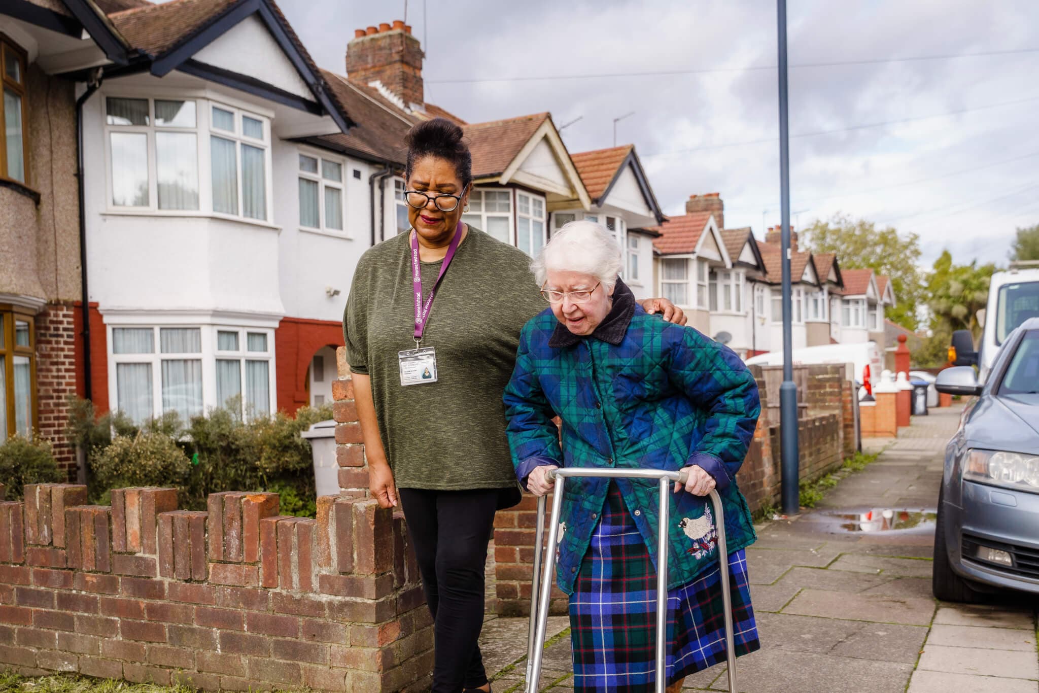A caregiver with a lanyard assists an elderly woman using a walker on a suburban sidewalk beside houses and parked cars. - Home Instead