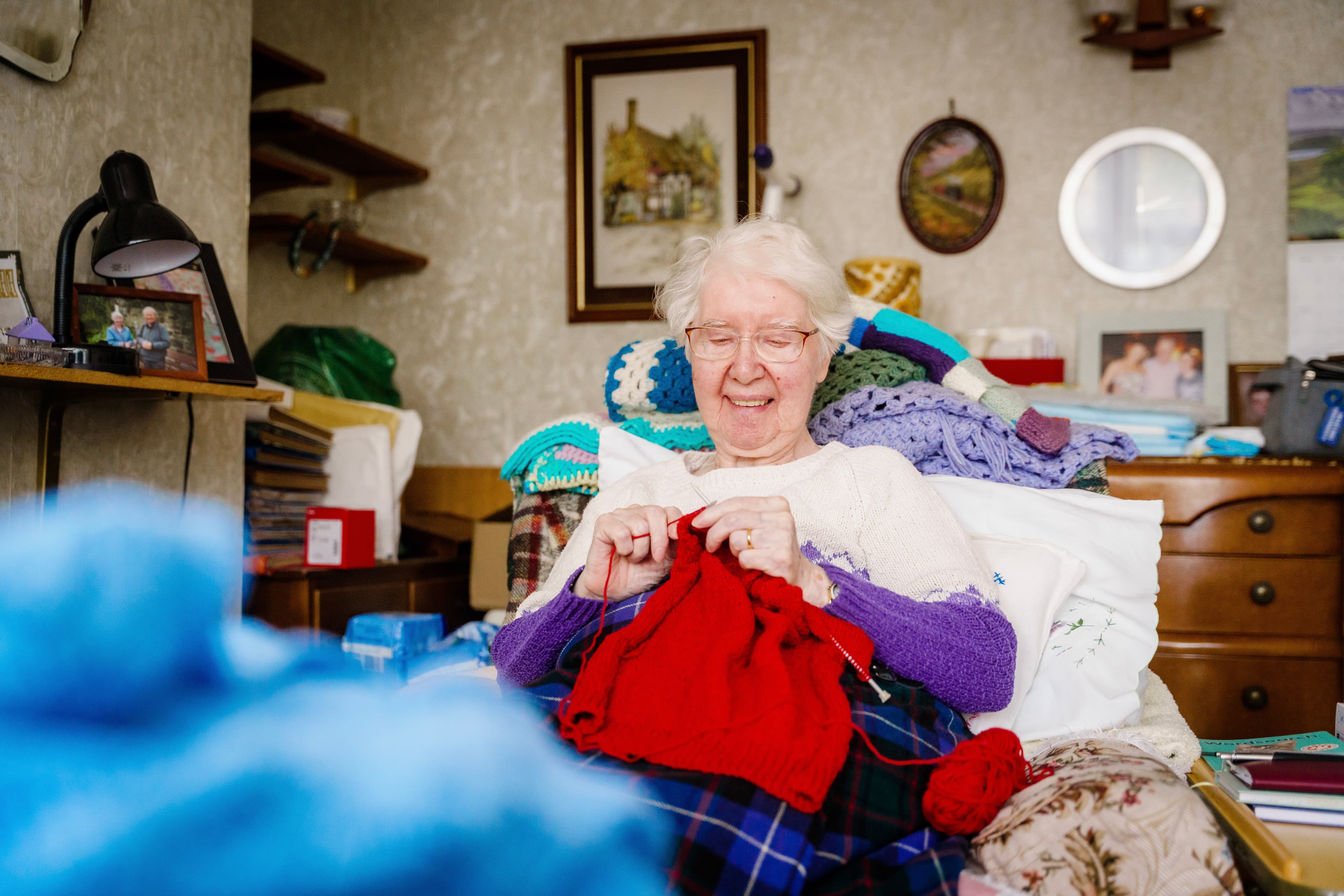 Elderly woman with glasses and white hair, smiling while knitting a red garment in a cozy room with shelves and pictures. - Home Instead