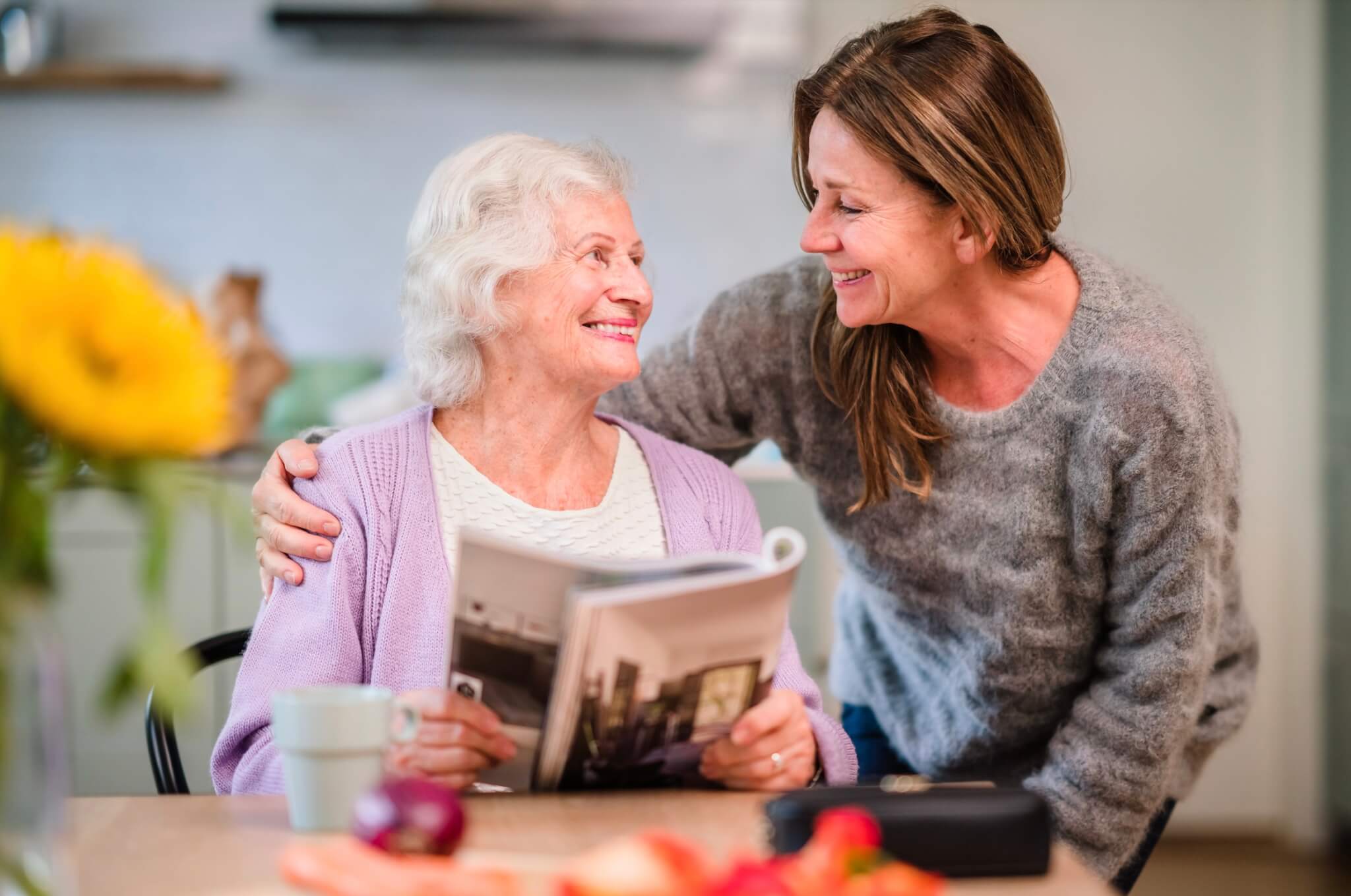 Older woman smiling, reading a magazine with a younger woman beside her, both appearing joyful in a cozy kitchen. - Home Instead
