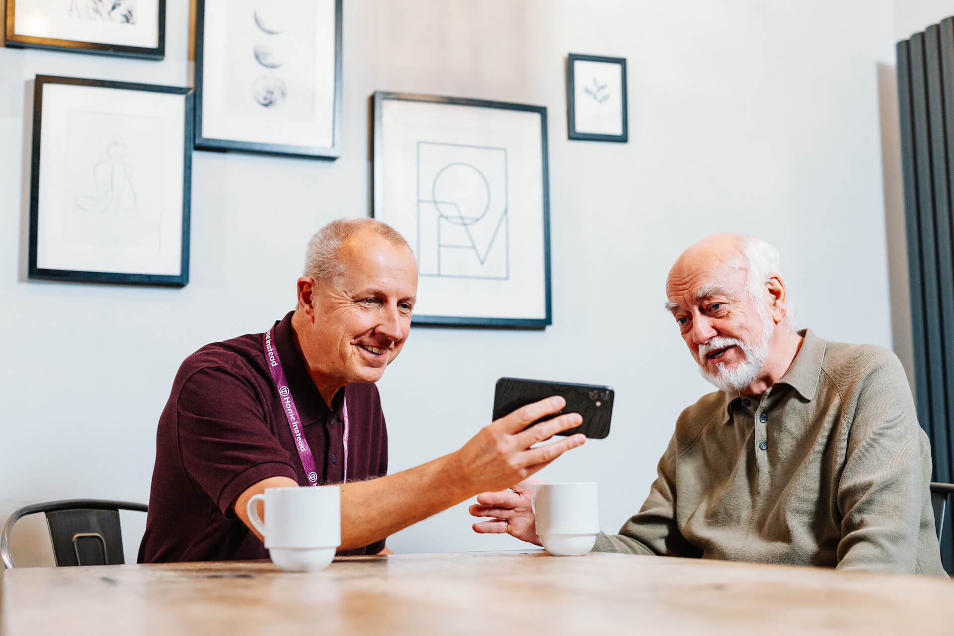 Two older men at a table, smiling as they look at a smartphone, with framed pictures on the wall behind them. - Home Instead