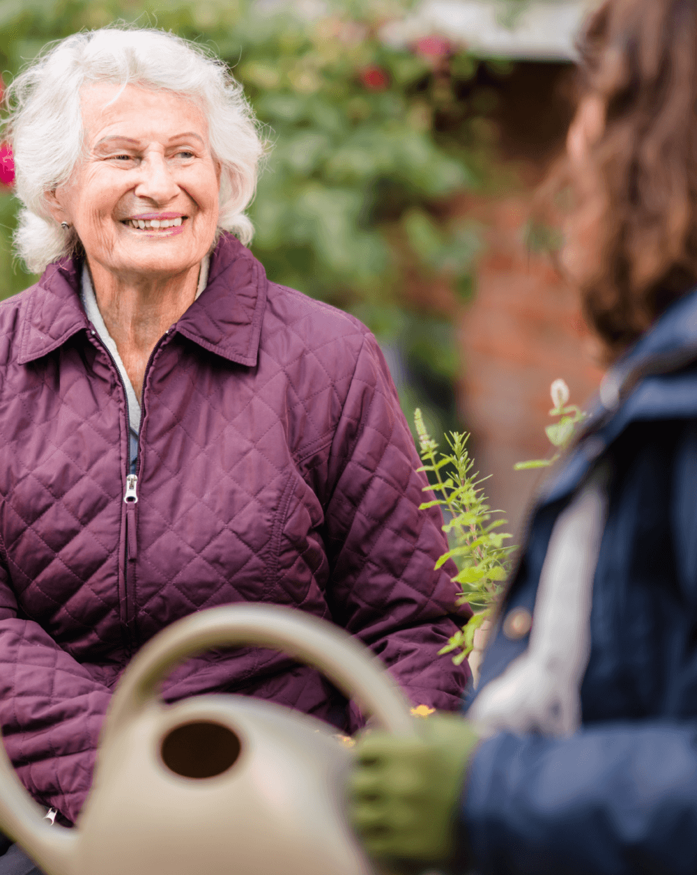 Elderly lady in a purple jacket smiles warmly while talking to another person in a garden setting, holding a watering can. - Home Instead