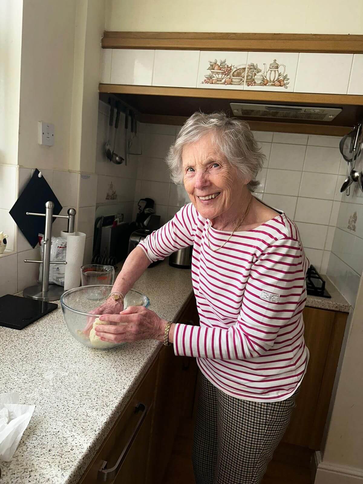 Elderly woman wearing a striped shirt kneads dough in a bowl on a kitchen counter, smiling at the camera. - Home Instead
