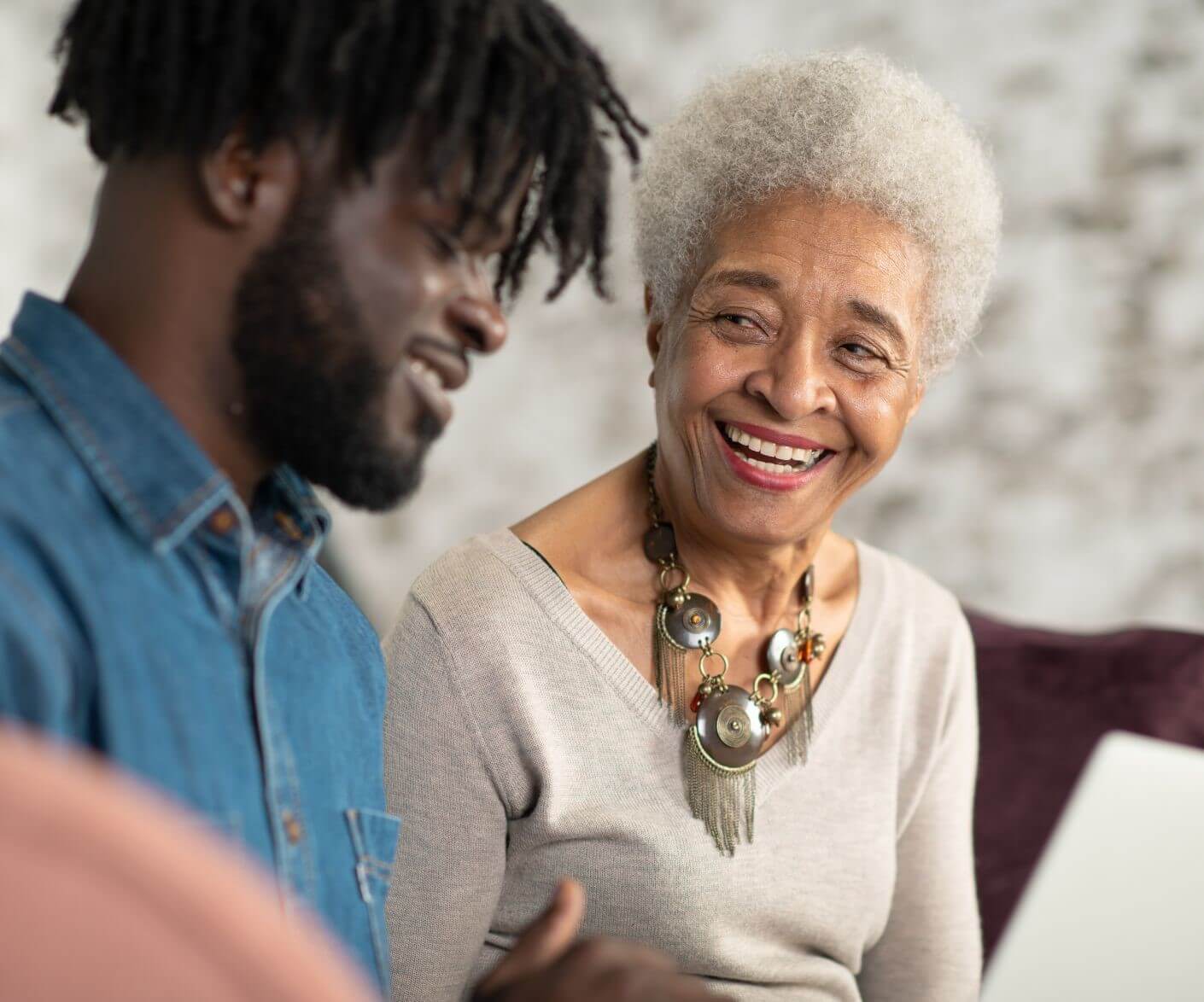 Older woman with white hair and a necklace smiling at a younger man with dreadlocks, both are seated and talking. - Home Instead