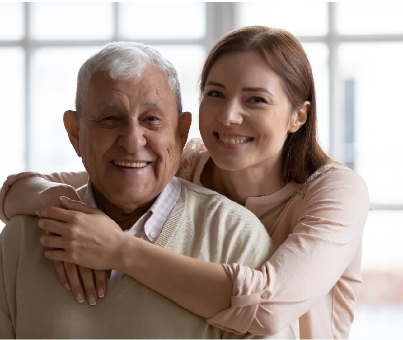 An older man and a young woman smile at the camera, with the woman hugging the man from behind. - Home Instead