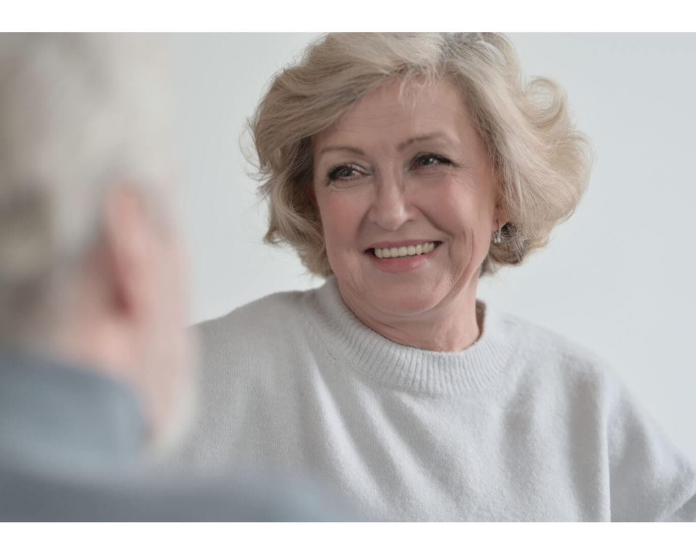 A smiling elderly woman with short, wavy hair looks at an elderly man with a blurred face in the foreground. - Home Instead