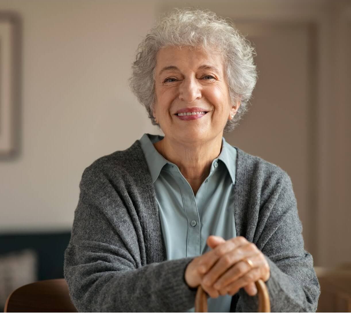 Elderly woman with short white hair and a warm smile, wearing a gray cardigan and holding a wooden cane indoors. - Home Instead