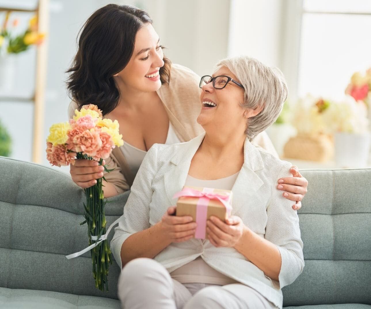 A woman gives an elderly woman flowers and a gift as they smile and sit on a sofa in a bright room. - Home Instead