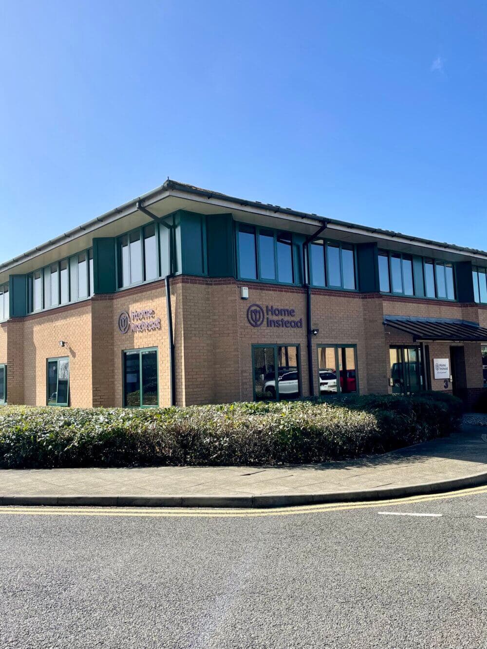 Two-story building with "Home Instead" signage on a clear day, surrounded by bushes and a curved driveway. - Home Instead