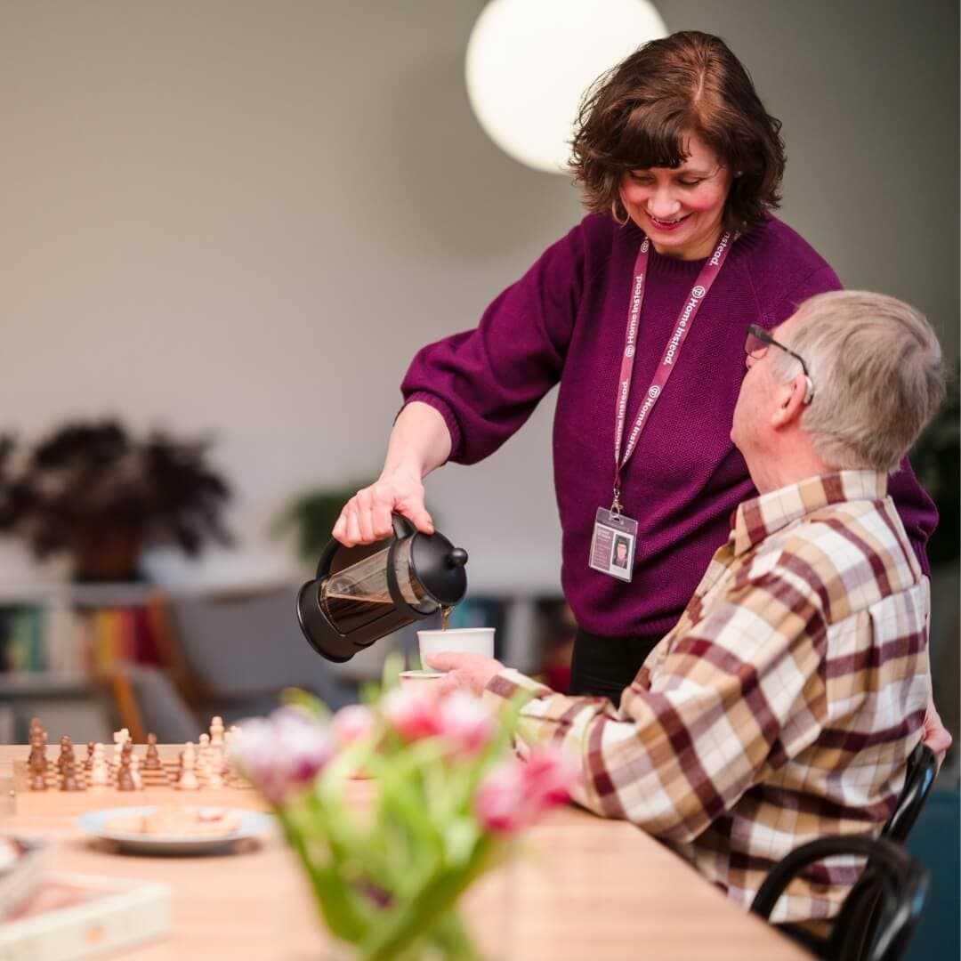 A woman in a purple sweater pours coffee for an elderly man seated at a table with a chessboard and flowers. - Home Instead