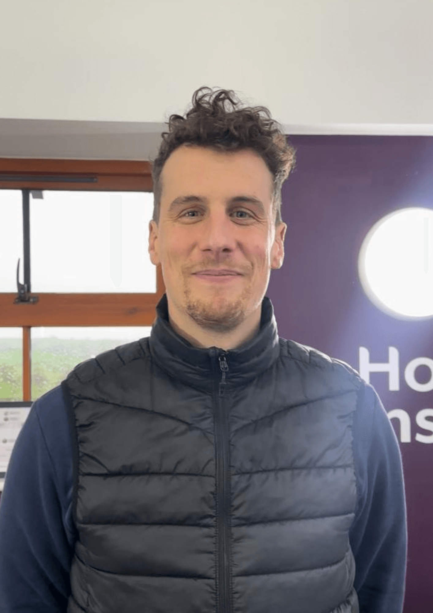 A man with curly hair, wearing a black puffer vest, smiles while standing indoors with a window in the background. - Home Instead