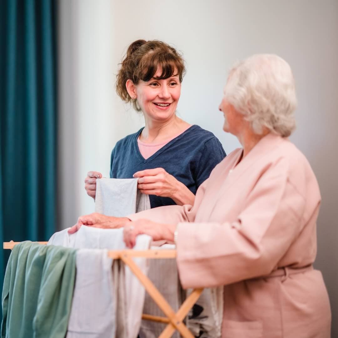 Two women smiling while folding laundry together; one holds a towel, the other stands near a drying rack. - Home Instead