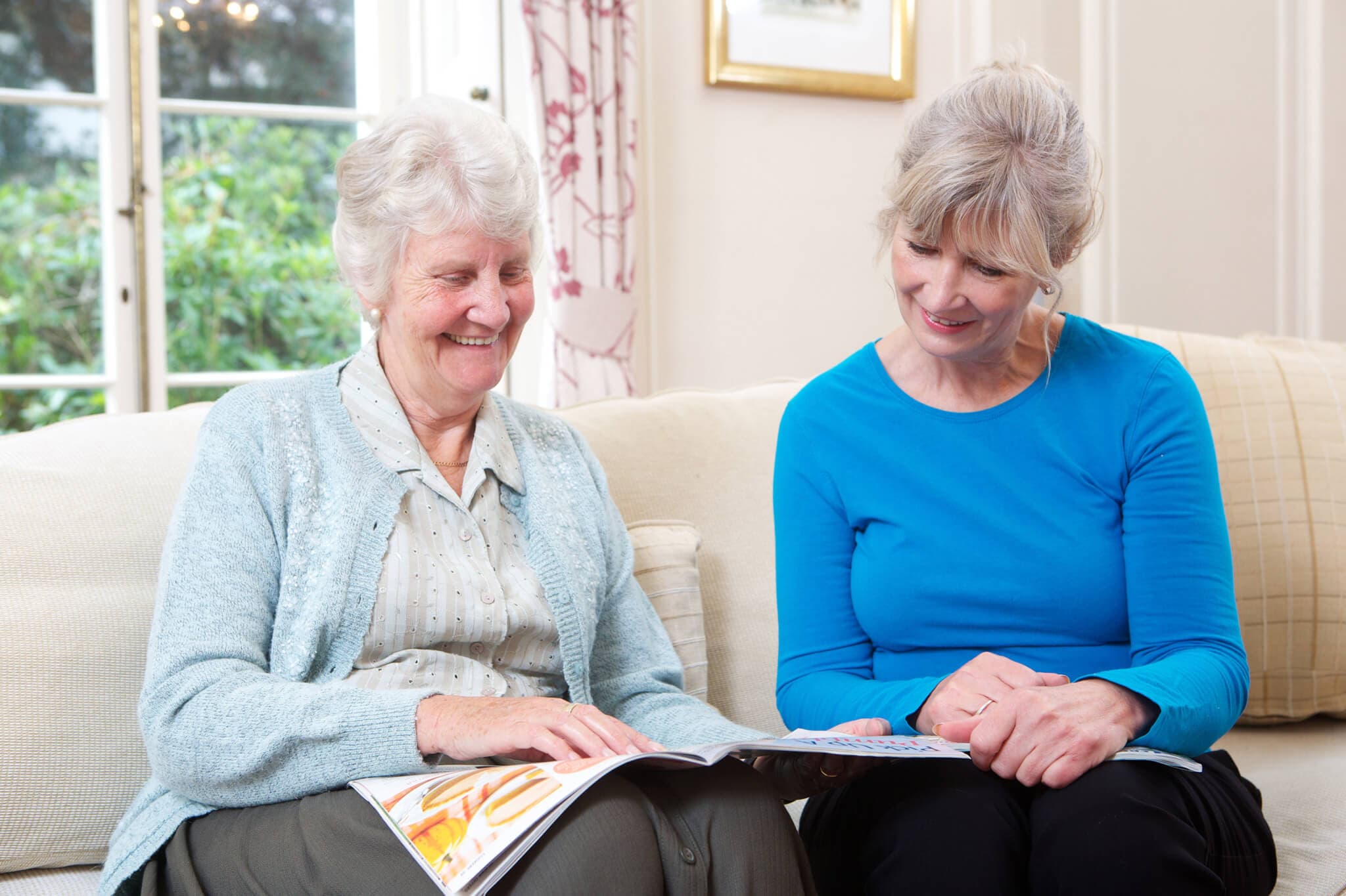 Two women sitting on a couch, smiling and reading a magazine together in a cozy, well-lit room. - Home Instead