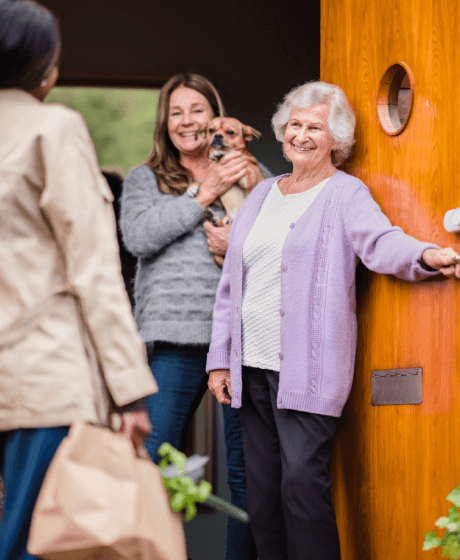 Two women greet a visitor at the door; one holds a dog while the other stands smiling in a purple sweater. - Home Instead