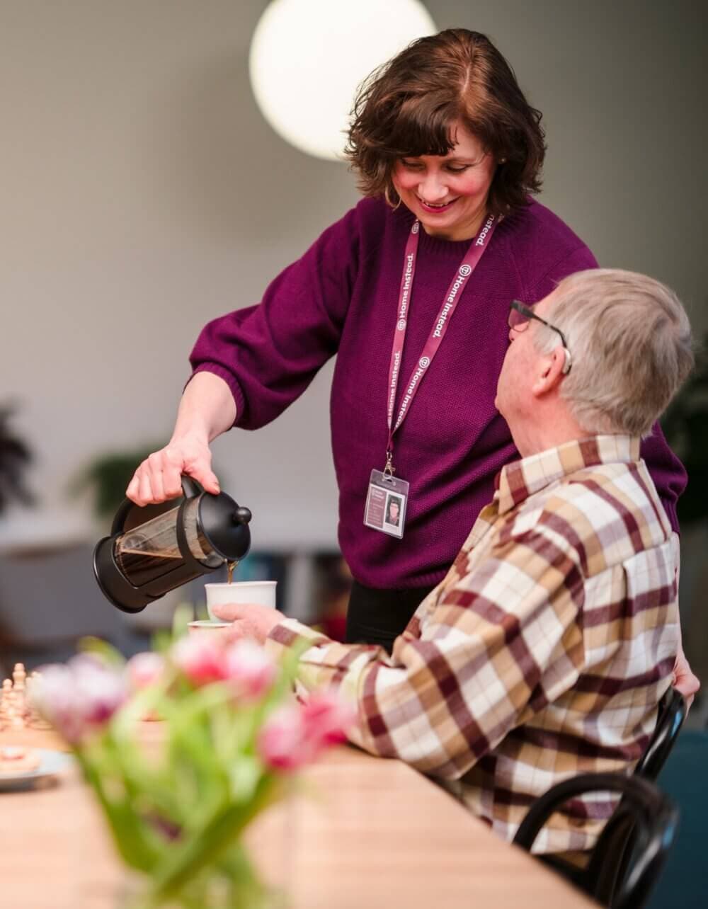 A woman pours coffee for an elderly man sitting at a table, with colorful flowers and a chessboard nearby. - Home Instead