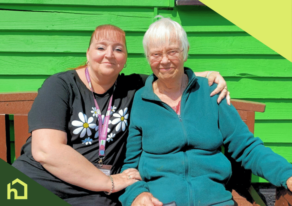 A younger woman and elderly woman sit smiling together on a bench in front of a bright green wall. - Home Instead