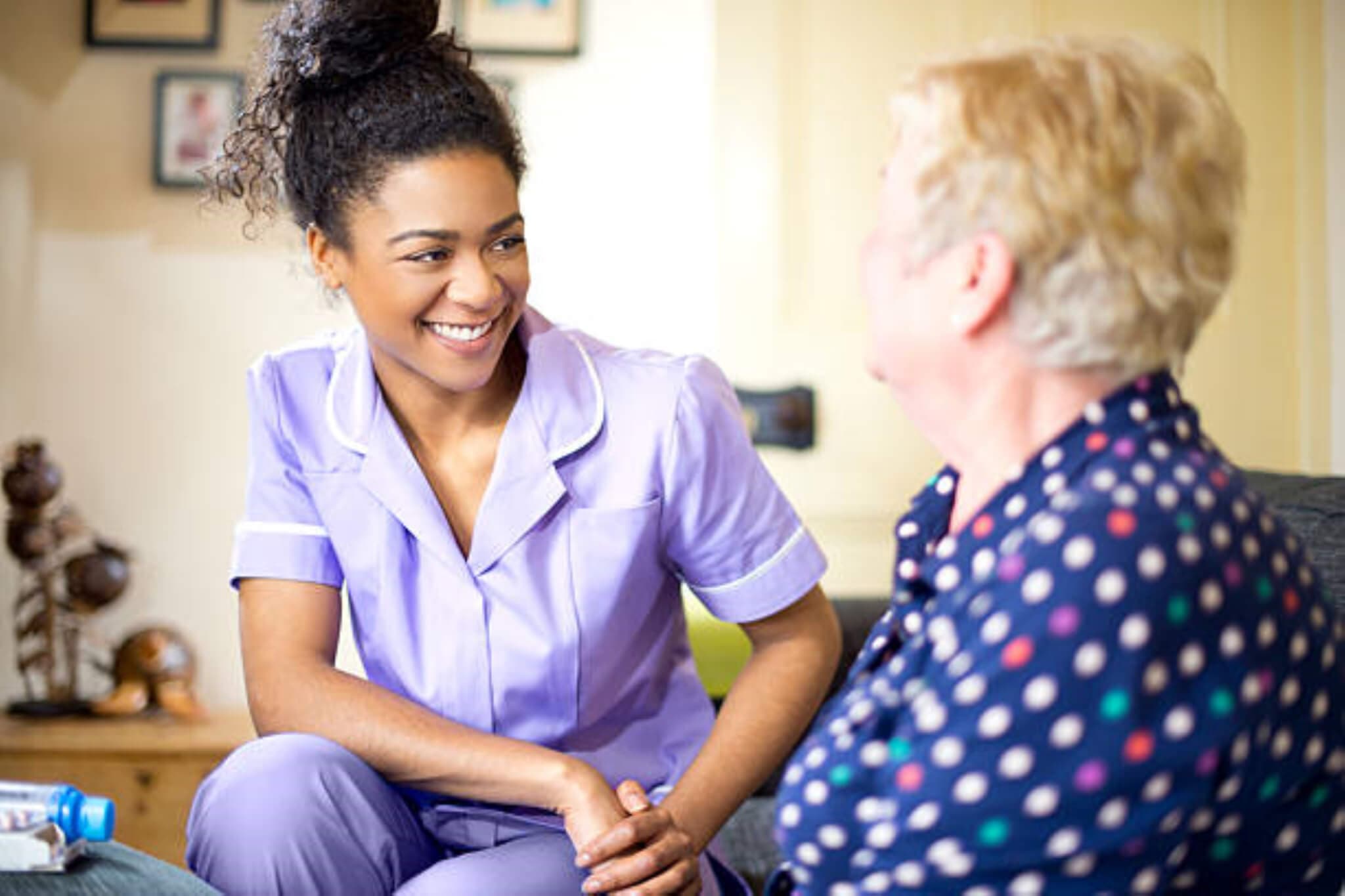 A smiling caregiver in a purple uniform interacting with an elderly person in a polka dot blouse. - Home Instead