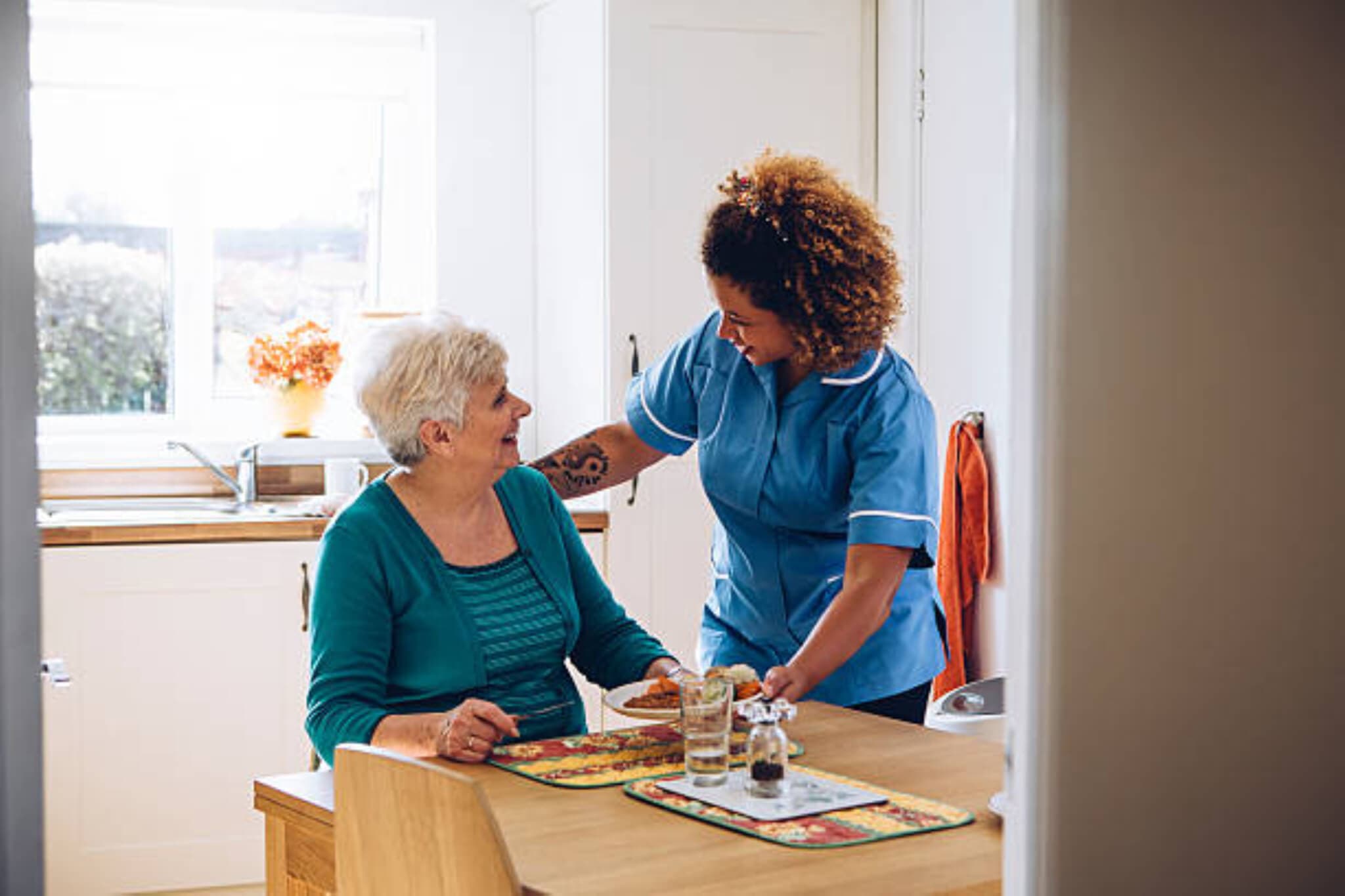 A caregiver in a blue uniform assists an elderly woman seated at a kitchen table, both smiling and conversing. - Home Instead