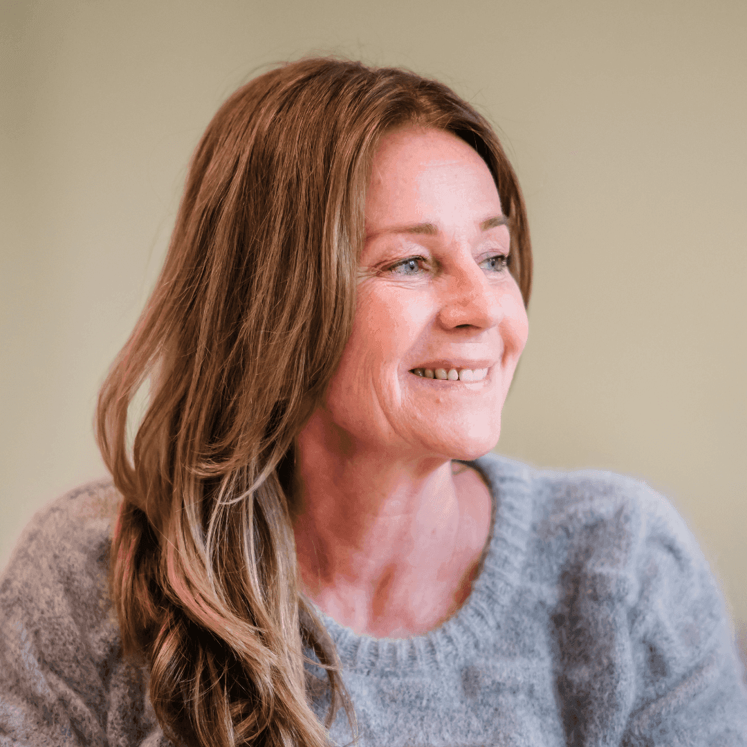 Smiling woman with long brown hair wearing a gray sweater, looking off to the side. - Home Instead