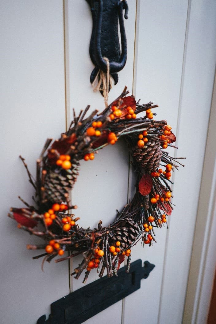 A wreath made of twigs, pinecones, and orange berries hangs on a white door. - Home Instead