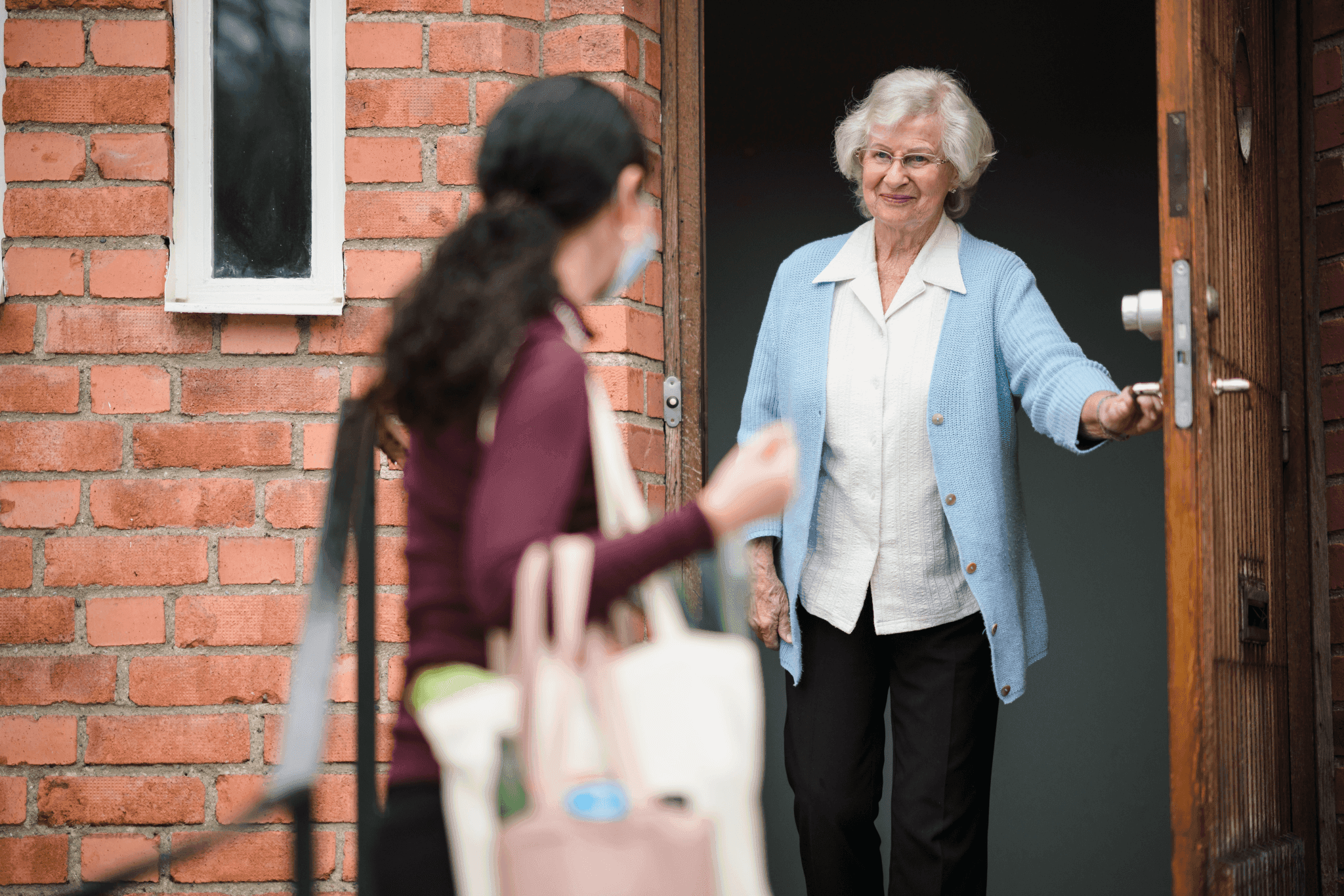 Elderly woman holding a door open with a smile as a younger woman with a bag walks towards the entrance. - Home Instead