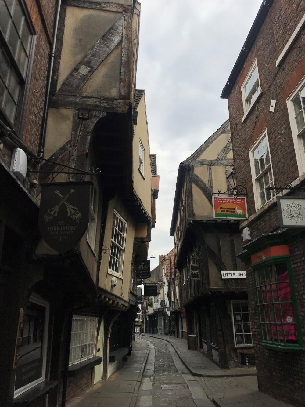 A narrow, cobblestone street lined with old, timber-framed buildings and shop signs under a cloudy sky. - Home Instead