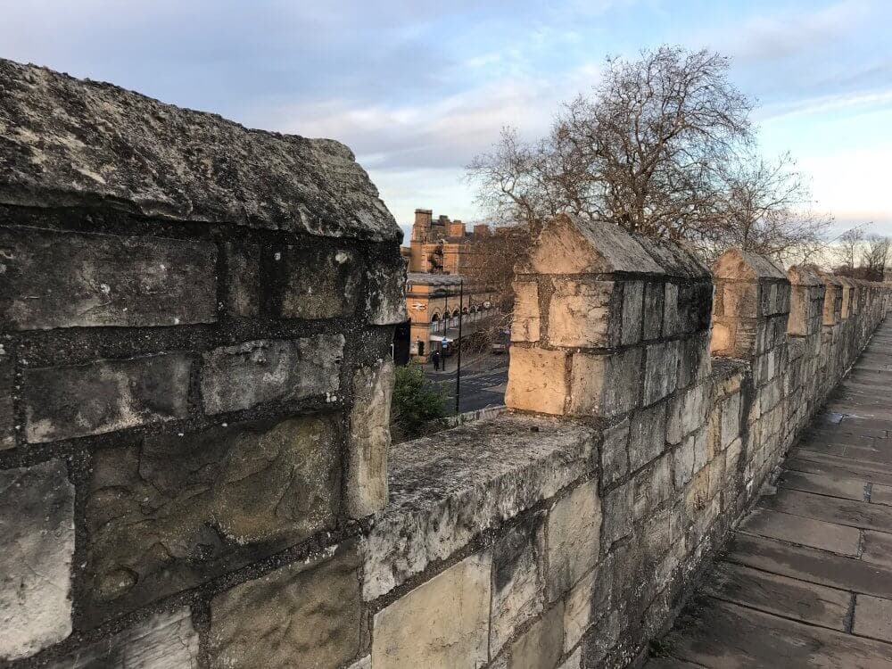 A weathered stone wall with crenellations overlooks a distant building and a tree under a partly cloudy sky. - Home Instead