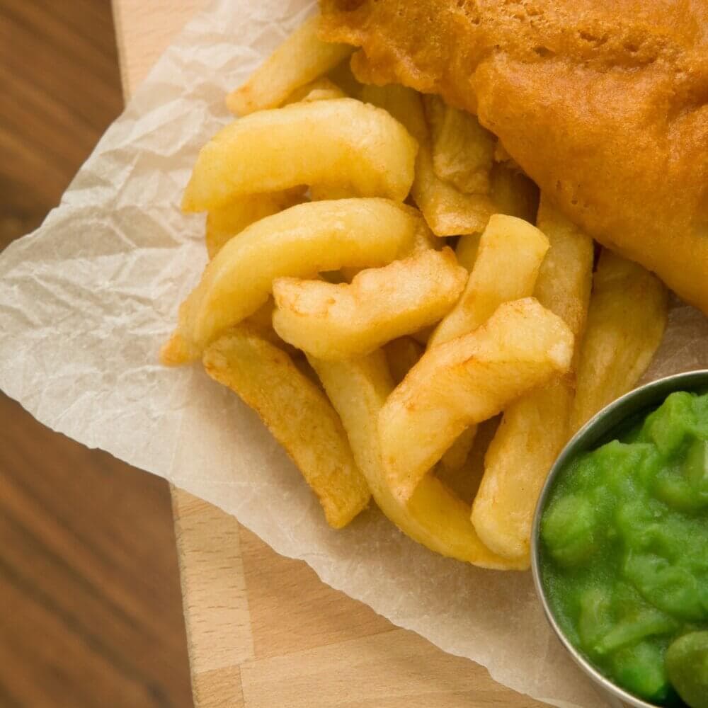 Close-up of crispy fries and a piece of battered fish, served with a small dish of mushy peas on a piece of parchment paper. - Home Instead