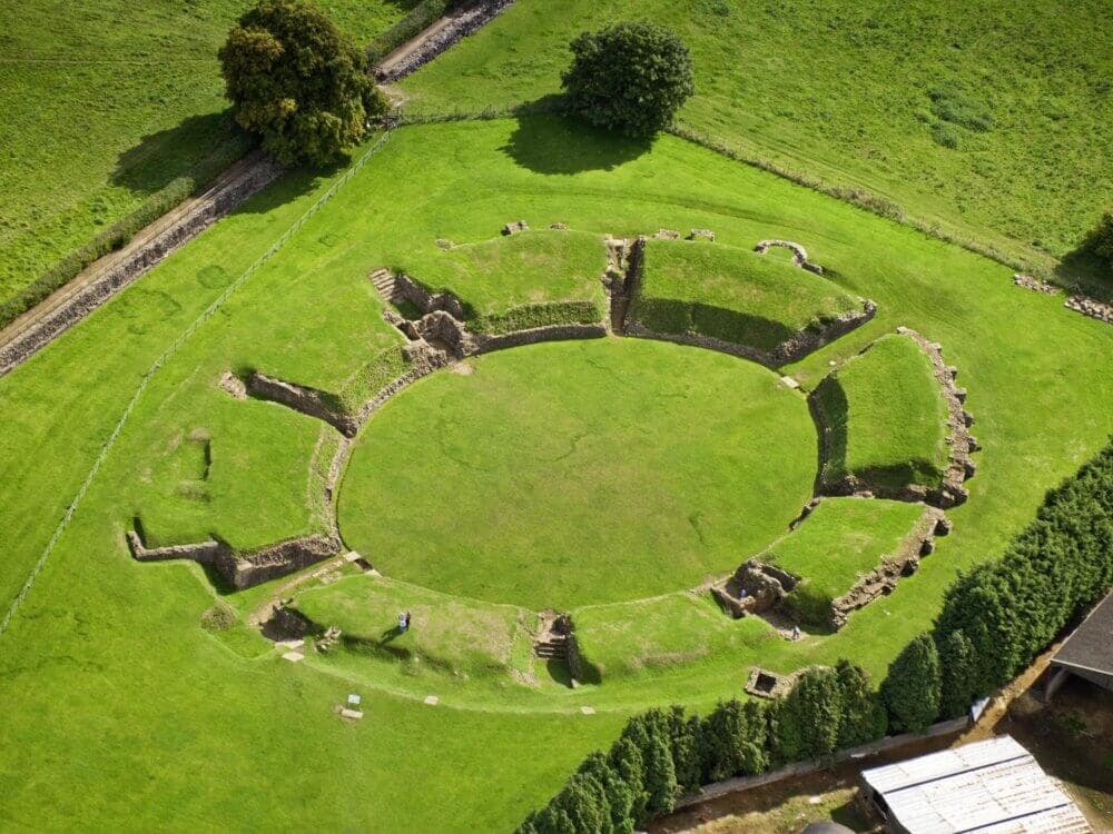 Aerial view of the ruins of a circular ancient amphitheater surrounded by green grass and trees. - Home Instead