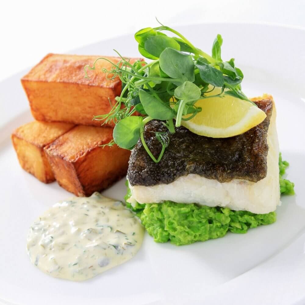 A plated dish with fish, pea puree, lemon wedge, greens, roasted potatoes, and a side of tartar sauce. - Home Instead